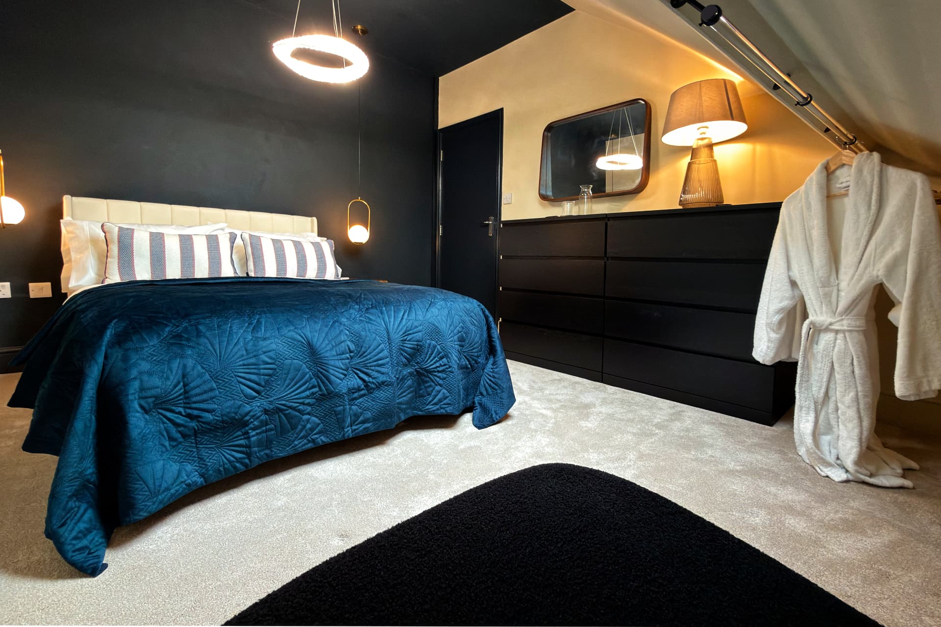Winter suite bedroom at a Soulful Stays serviced apartment in Darlington, with a king bed dressed in a deep teal velvet throw, dark walls, a ring pendant light, warm bedside lamp and a white robe hanging beside the drawers